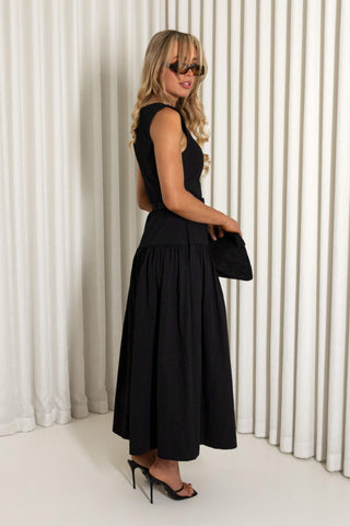 A woman modeling a sleeveless black midi dress with a gathered waist and flared skirt, styled with black high heels, sunglasses, and a black clutch, standing against a white curtain backdrop.