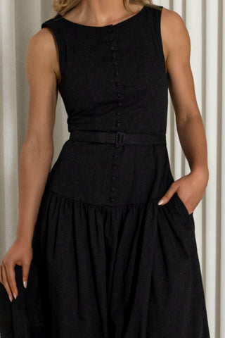 A woman wearing a sleeveless black midi dress with a buttoned front, matching belt, and side pockets, photographed against a neutral curtain background.