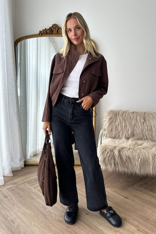 A woman stands in a room wearing the Eric brown jacket over a white top, dark wide-leg jeans, and black loafers, while holding a matching brown tote bag. A large ornate mirror and a fuzzy chair are visible in the background.