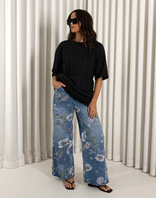 Woman wearing a black top and blue floral jeans standing against a white curtain background. High waisted wide leg denim jeans with floral print