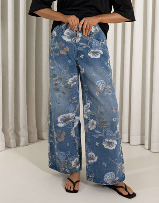 Woman wearing a black top and blue floral jeans standing against a white curtain background. High waisted wide leg denim jeans with floral print