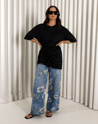 Woman wearing a black top and blue floral jeans standing against a white curtain background. High waisted wide leg denim jeans with floral print