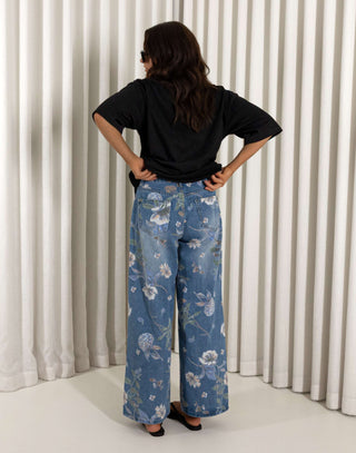 Woman wearing a black top and blue floral jeans standing against a white curtain background. High waisted wide leg denim jeans with floral print