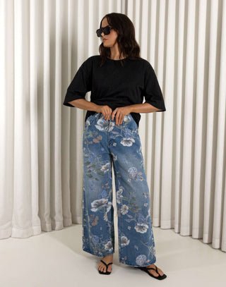 Woman wearing a black top and blue floral jeans standing against a white curtain background. High waisted wide leg denim jeans with floral print