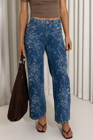 A model wearing blue floral print wide-leg cropped jeans, paired with a brown crop top, black sandals, and holding a brown tote bag, set against a background of white curtains.