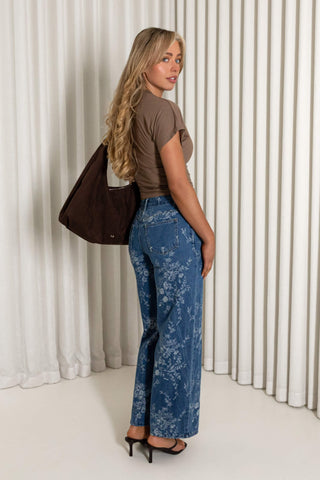 A woman stands in profile wearing blue floral wide-leg pants, a brown short-sleeve top, and black heels, while carrying a dark brown shoulder bag against a white pleated curtain backdrop.
