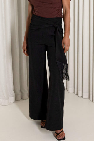A model wearing black wide-leg wrap pants with a side tie and fringe detail, paired with a brown top and black sandals, standing in front of white curtains.