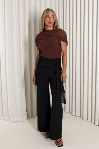 A woman models a dark brown draped short-sleeve top paired with black wide-leg trousers featuring a side tie and fringe detail, set against a background of white vertical drapes.