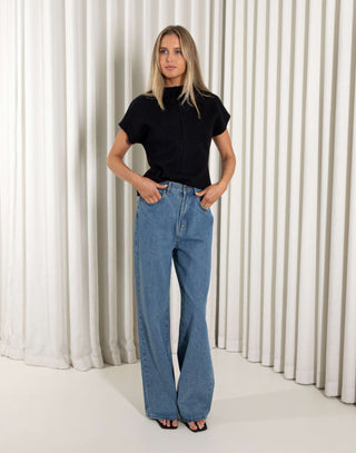 Woman wearing a black knit top and blue jeans standing against a white curtain background