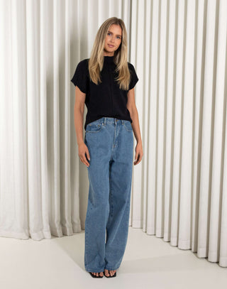 Woman wearing a black knit top and blue jeans standing against a white curtain background