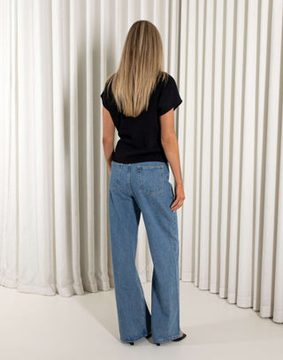 Woman wearing a black knit top and blue jeans standing against a white curtain background