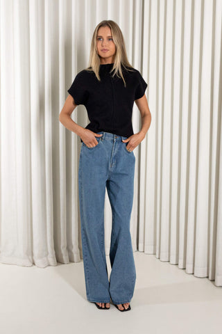 A woman modeling a black short-sleeve mock neck top and wide-leg blue denim jeans, standing against a white curtain backdrop.