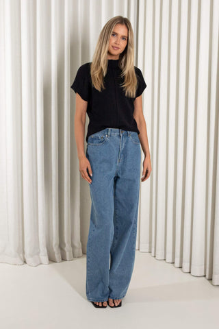 A woman modeling a black short-sleeve top and light blue wide-leg jeans against a white curtain backdrop.