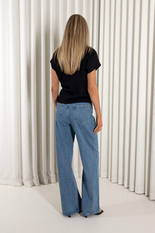 A woman viewed from behind wearing a black short-sleeve t-shirt and light blue wide-leg jeans, standing in front of a white curtain backdrop.