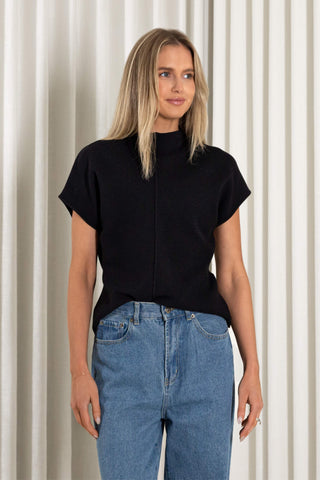 A woman modeling a black short-sleeve mock neck knit top with a front zipper, paired with blue jeans, standing against a cream-colored pleated curtain backdrop.