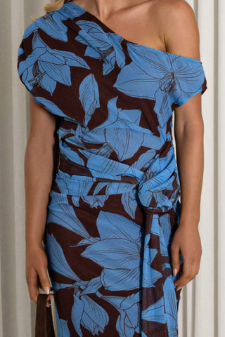 A woman wearing a one-shoulder wrap dress with a bold blue and dark brown floral print, standing against a neutral curtain background.