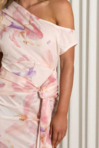 A close-up of a woman wearing a one-shoulder wrap dress with a pink and purple watercolor floral print on a light cream base, styled with a matching tie belt at the waist.