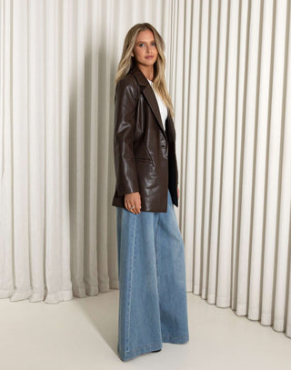 Woman wearing a brown leather jacket and blue jeans against a white curtain background