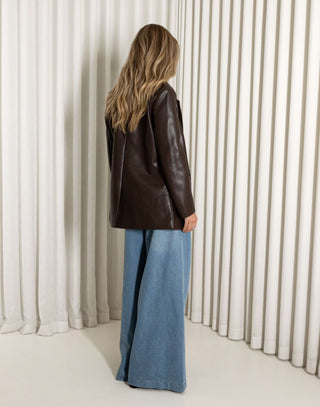 Woman wearing a brown leather jacket and blue jeans against a white curtain background