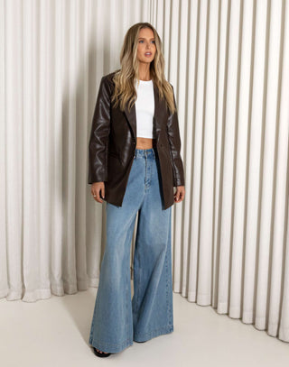 Woman wearing a brown leather jacket and blue jeans against a white curtain background