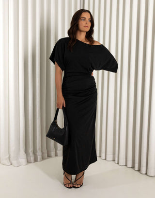Woman wearing a black off-shoulder dress against a white curtain background. Black off shoulder midi dress with batwing sleeves and gathered sides
