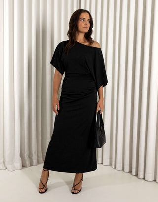 Woman wearing a black off-shoulder dress against a white curtain background. Black off shoulder midi dress with batwing sleeves and gathered sides