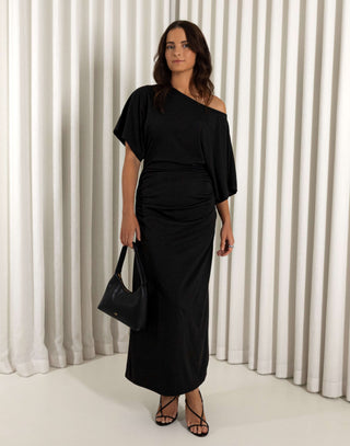 Woman wearing a black off-shoulder dress against a white curtain background. Black off shoulder midi dress with batwing sleeves and gathered sides