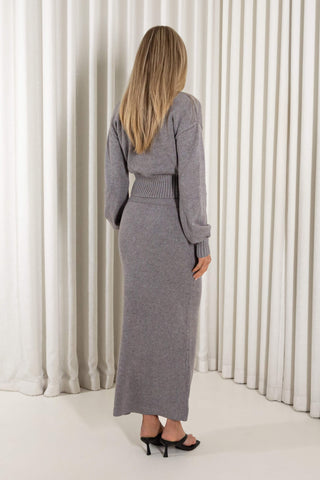 A woman with long blonde hair is shown from the back wearing a gray knit long-sleeve maxi dress with a ribbed waistband, standing in front of a white pleated curtain backdrop.