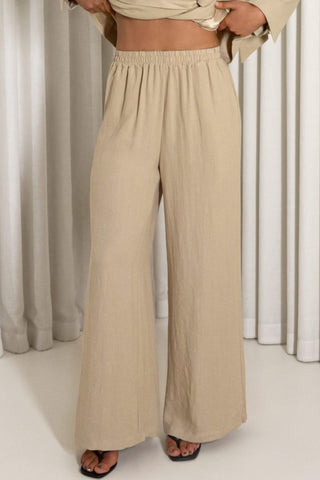 A person wearing loose, wide-leg beige pants with an elastic waistband, paired with a matching beige cropped top and black sandals, standing in front of white curtains.