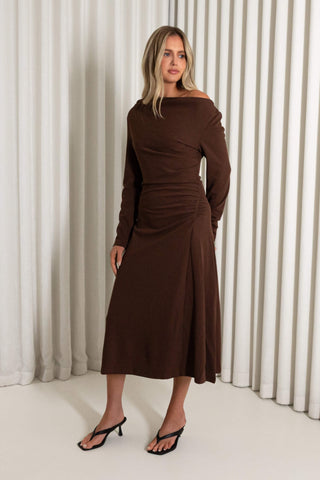 A woman modeling a dark brown off-the-shoulder midi dress with long sleeves and gathered draping, paired with black heeled sandals, set against a background of vertical white drapes.