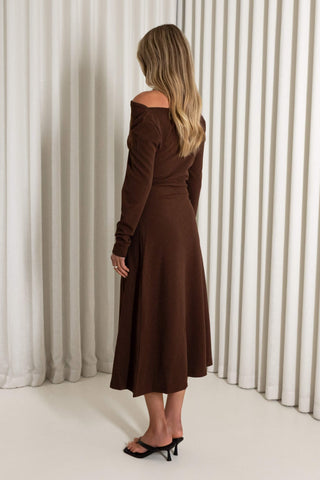 A woman stands with her back to the camera wearing a long-sleeve off-the-shoulder brown midi dress and black mule heels, posed in front of white vertical drapes.
