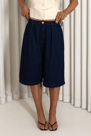 A model wearing dark blue denim culottes with a pleated front, paired with a cream sleeveless top and black thong sandals, standing in front of white pleated curtains.