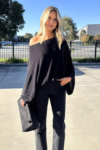 A woman wearing a black off-the-shoulder knit sweater and black pants, holding a black clutch bag, standing outdoors in a sunny parking lot.