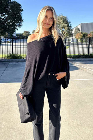 A woman wearing a black off-the-shoulder knit sweater and black pants, holding a black clutch bag, standing outdoors in a sunny parking lot.