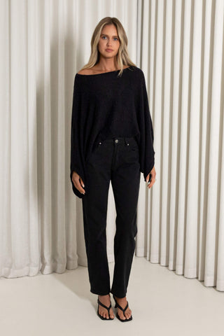 A woman modeling a black off-the-shoulder knit sweater and black straight-leg jeans against a background of white vertical drapes.