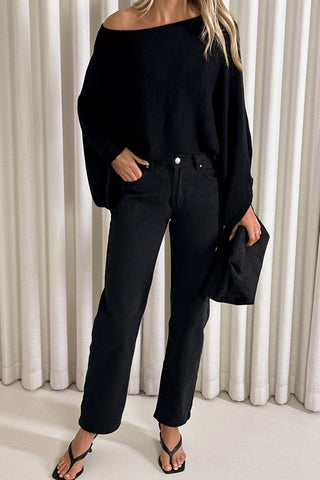 A woman wearing a black off-the-shoulder knit sweater and black straight-leg jeans, holding a black clutch bag, standing in front of white pleated curtains.