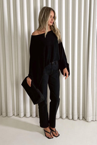 A woman modeling a black off-the-shoulder knit top with long balloon sleeves, paired with black jeans and black sandals, standing in front of white pleated curtains.