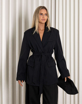 Woman wearing a black tie blazer and black pants standing against a white curtain background