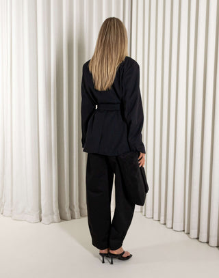 Woman wearing a black tie blazer and black pants standing against a white curtain background