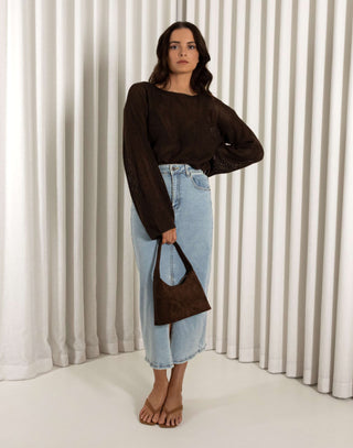 Woman wearing a brown top and blue denim skirt holding a brown bag against a white curtain background Brown open weave knit long sleeve top