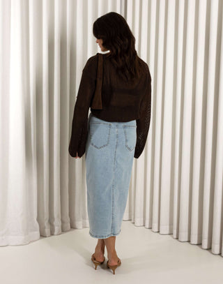Woman wearing a brown top and blue denim skirt holding a brown bag against a white curtain background Brown open weave knit long sleeve top