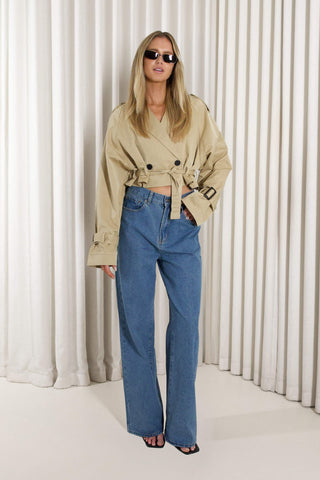 A woman modeling a beige double-breasted belted trench coat and high-waisted blue wide-leg jeans, standing against a white pleated curtain backdrop.