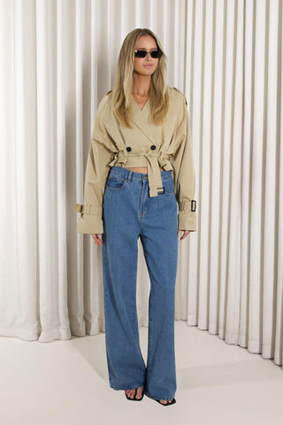 A woman modeling a beige double-breasted belted trench coat and high-waisted blue wide-leg jeans, standing against a white pleated curtain backdrop.