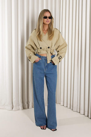 A woman modeling a beige double-breasted belted trench coat and high-waisted blue wide-leg jeans, standing against a white pleated curtain backdrop.