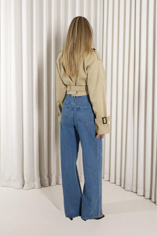 A woman modeling a beige double-breasted belted trench coat and high-waisted blue wide-leg jeans, standing against a white pleated curtain backdrop.