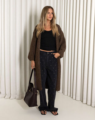 Woman wearing a brown cardigan, black top, and dark pants with white patterns, holding a brown bag against a white curtain background.