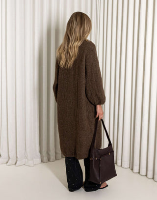 Person wearing a long brown coat holding a matching handbag against a white curtain background