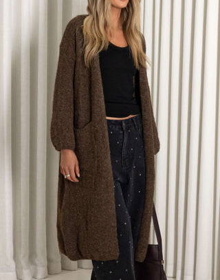 Person wearing a long brown cardigan over a black top and jeans, standing against a white curtain background.