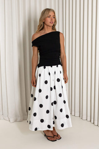 A woman modeling a black off-the-shoulder top paired with a white midi skirt featuring large black polka dots, standing in front of a white curtain backdrop.