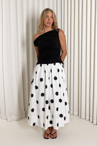 A woman modeling a black one-shoulder top paired with a white midi skirt featuring large black polka dots, standing in front of white pleated curtains.